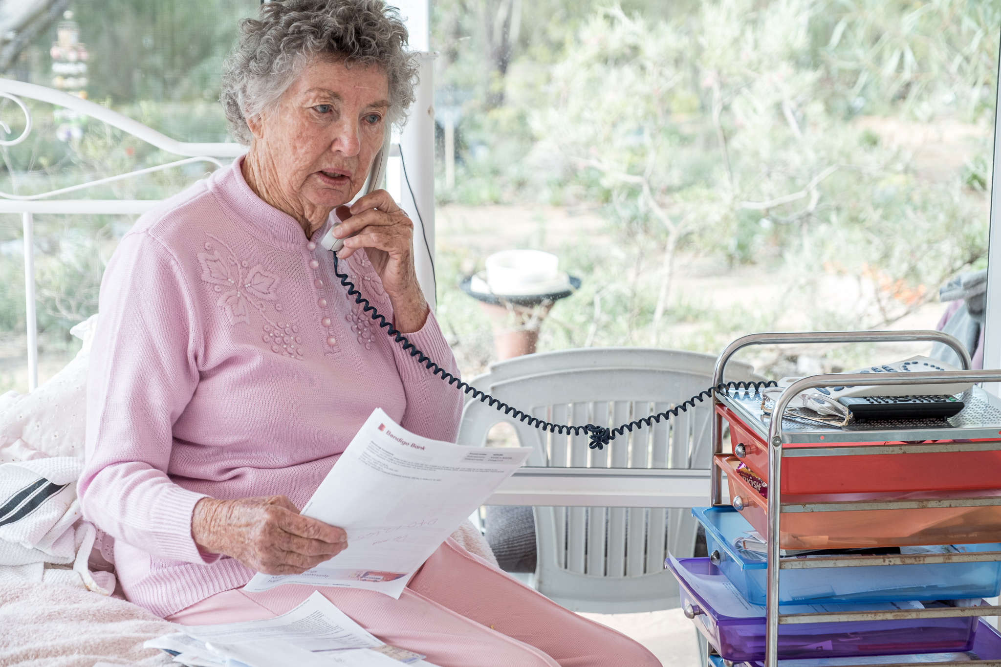 Older woman speaks on the phone by the window holding paperwork